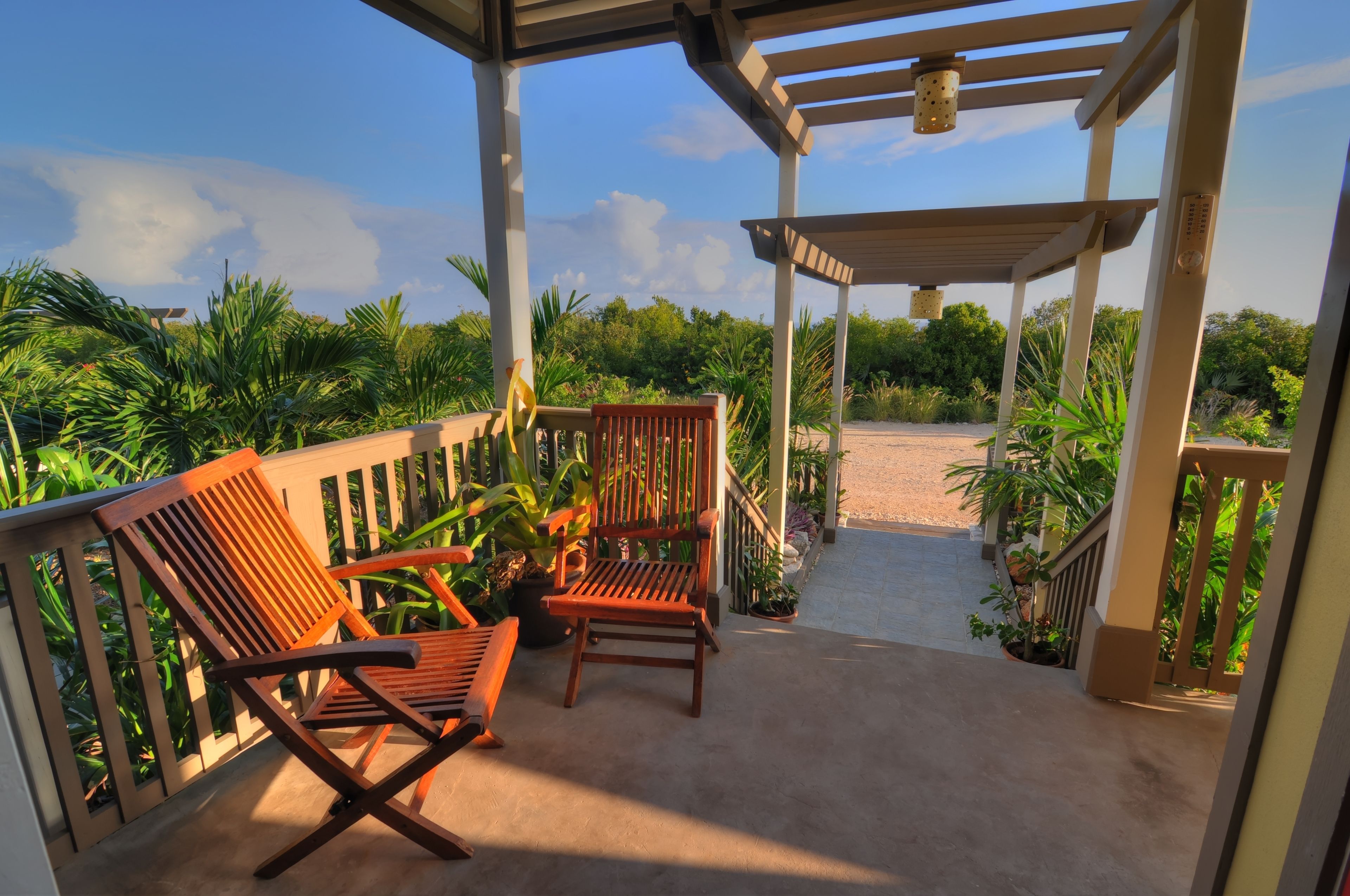 A covered outdoor seating area with wooden chairs and a pergola, overlooking a garden with various plants and a pathway.
