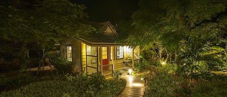 A small, illuminated house surrounded by lush greenery and a wooden pathway leading to it.