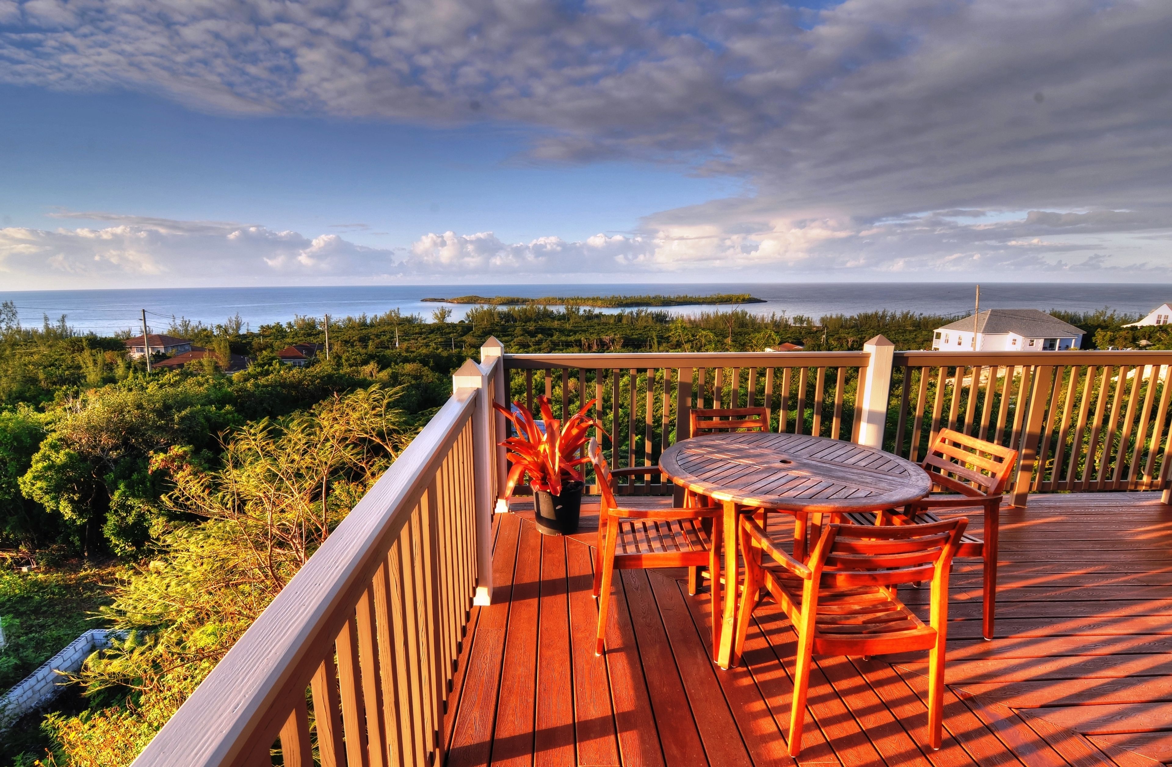 Una terraza de madera con una mesa redonda y sillas, con vista a un paisaje costero e islas distantes.