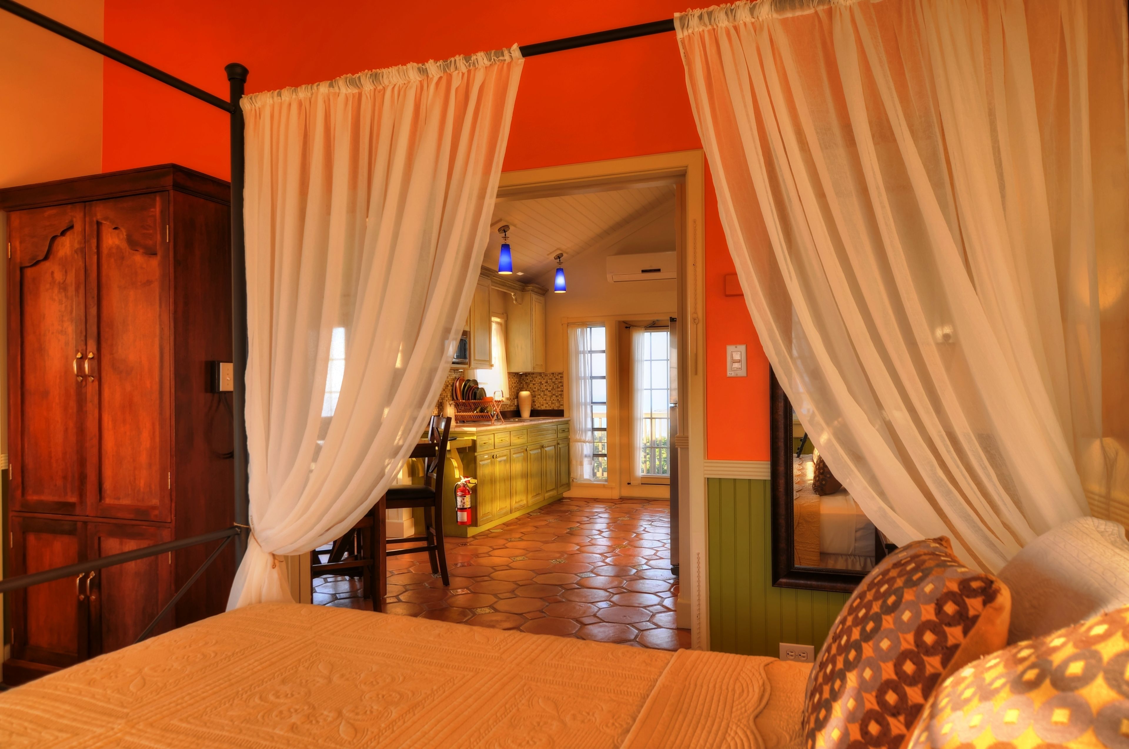A bedroom with a four-poster bed, a wooden wardrobe, and a kitchen area visible through an open door.