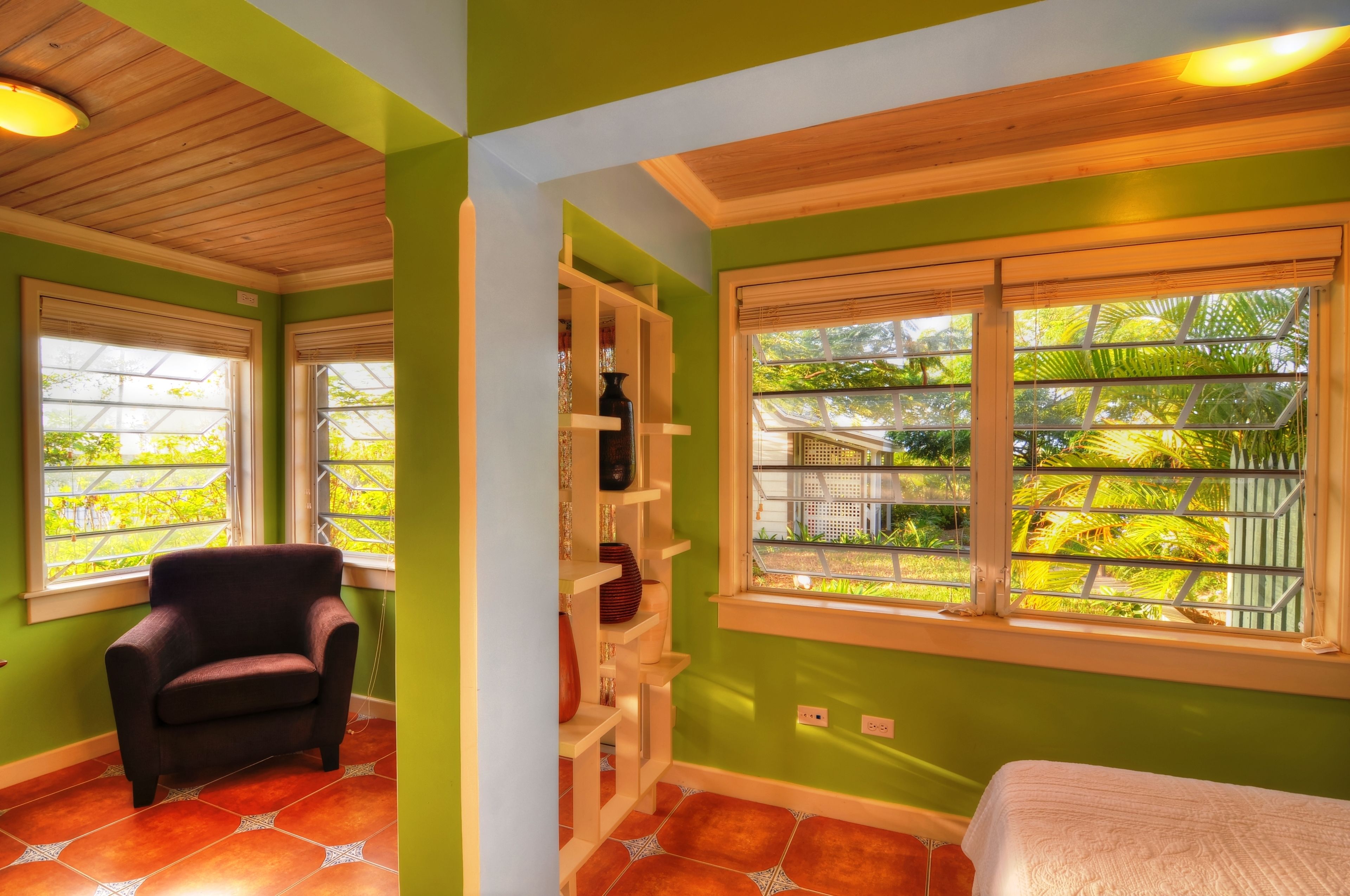 Une pièce aux murs verts, au plafond en bois et dotée d’une fenêtre donnant sur un paysage verdoyant. On y trouve une chaise et une étagère sur laquelle sont disposés des objets décoratifs.