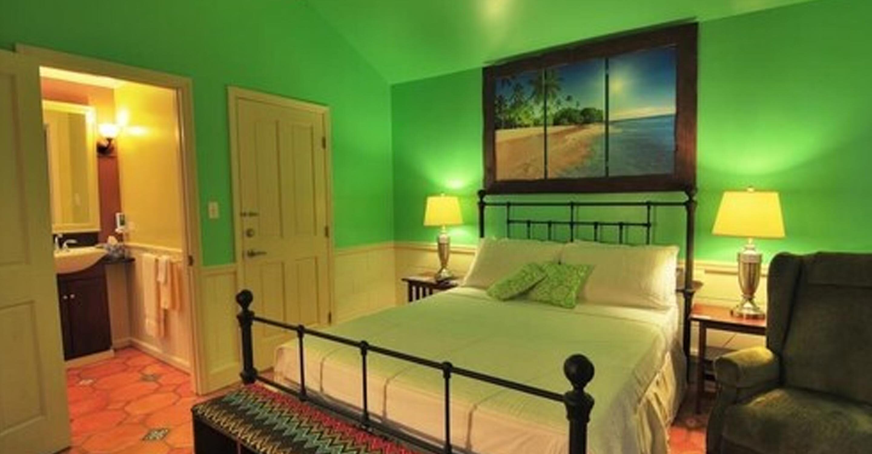 A room with a bed, two bedside lamps, a painting above the bed, and a chair.