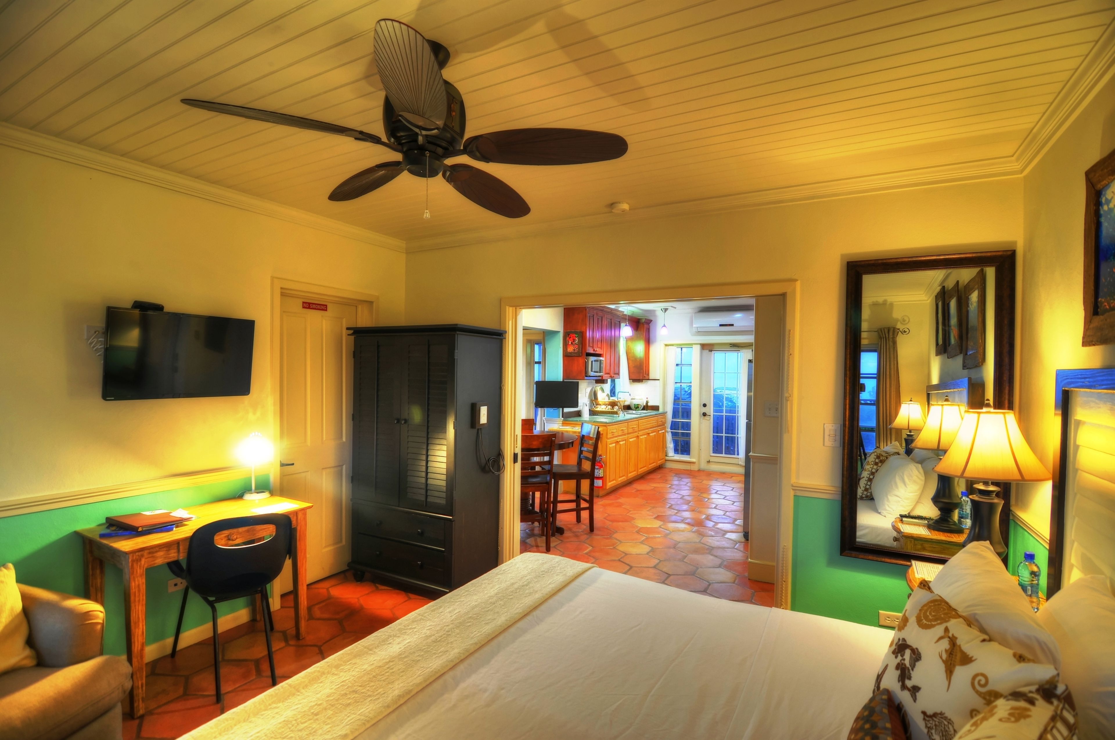 A hotel room with a bed, desk, chair, and a ceiling fan. There is a view of a kitchen and dining area through an open door.