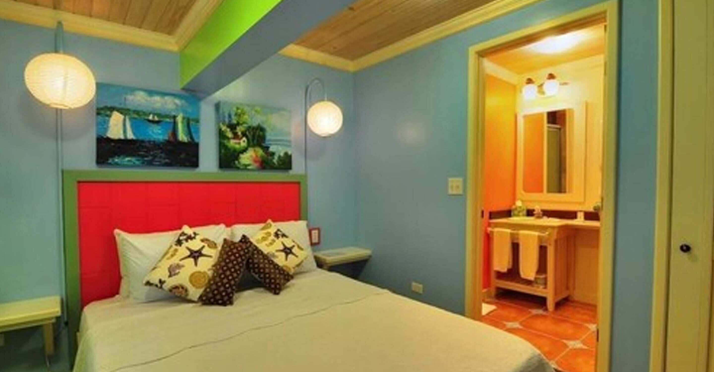 A bedroom with a bed, a red headboard, two wall-mounted lights, and a painting above the bed.