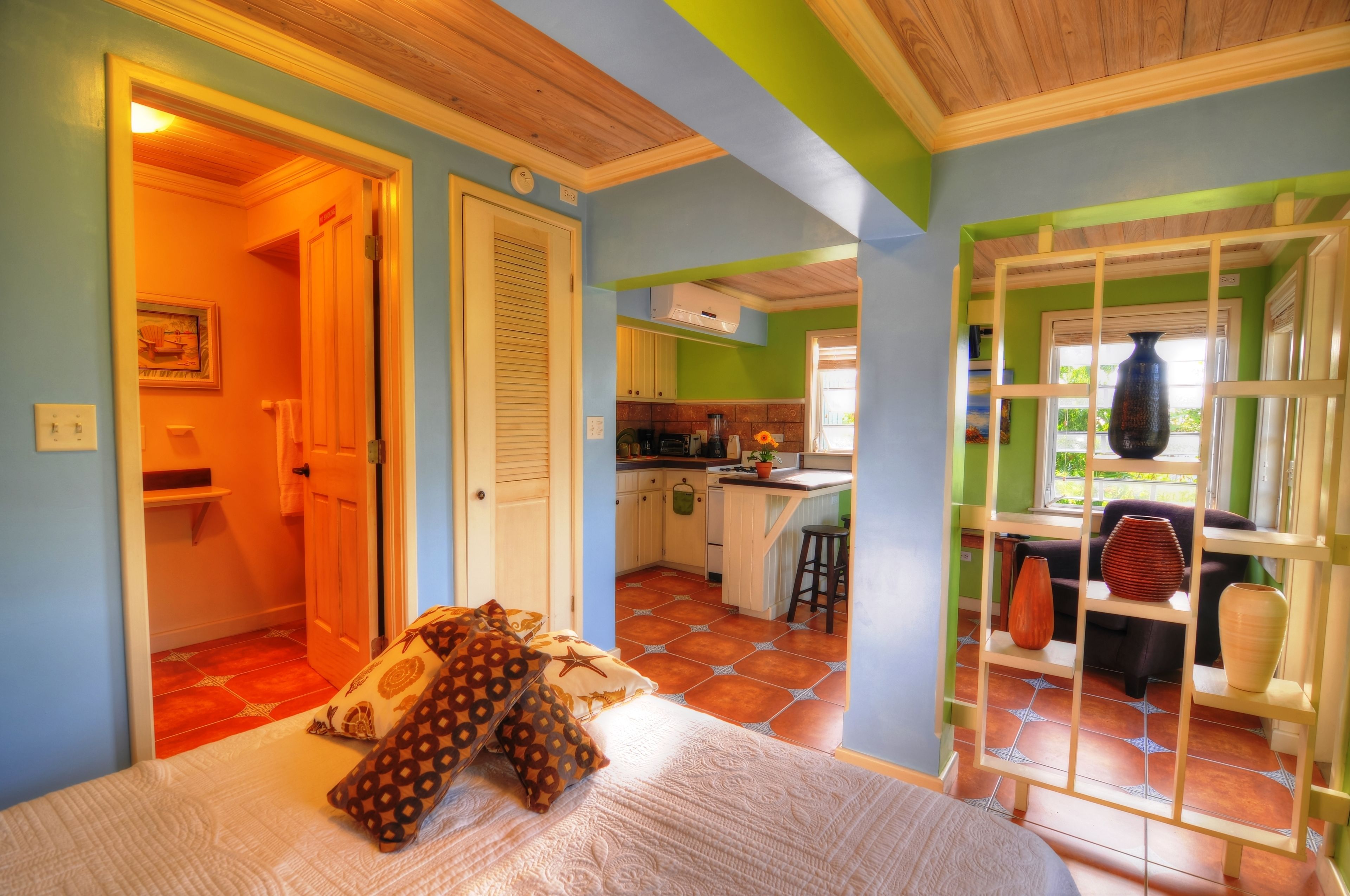 A bedroom with a bed, pillows, and a view into a kitchen and dining area.