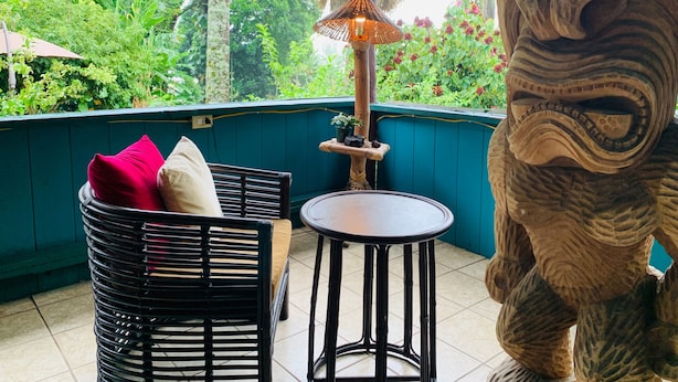 Una habitación con vistas a un follaje exuberante, un sofá de mimbre negro con cojines rojos y blancos, una pequeña mesa redonda y una escultura decorativa de piedra.