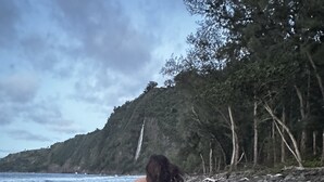 Una persona caminando sobre una playa rocosa con una colina boscosa al fondo.