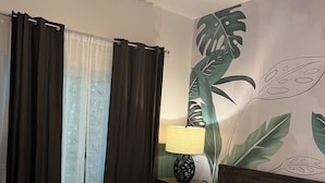 Habitación de hotel con cama, cortinas, mesita de noche con lámpara y un mural en la pared.