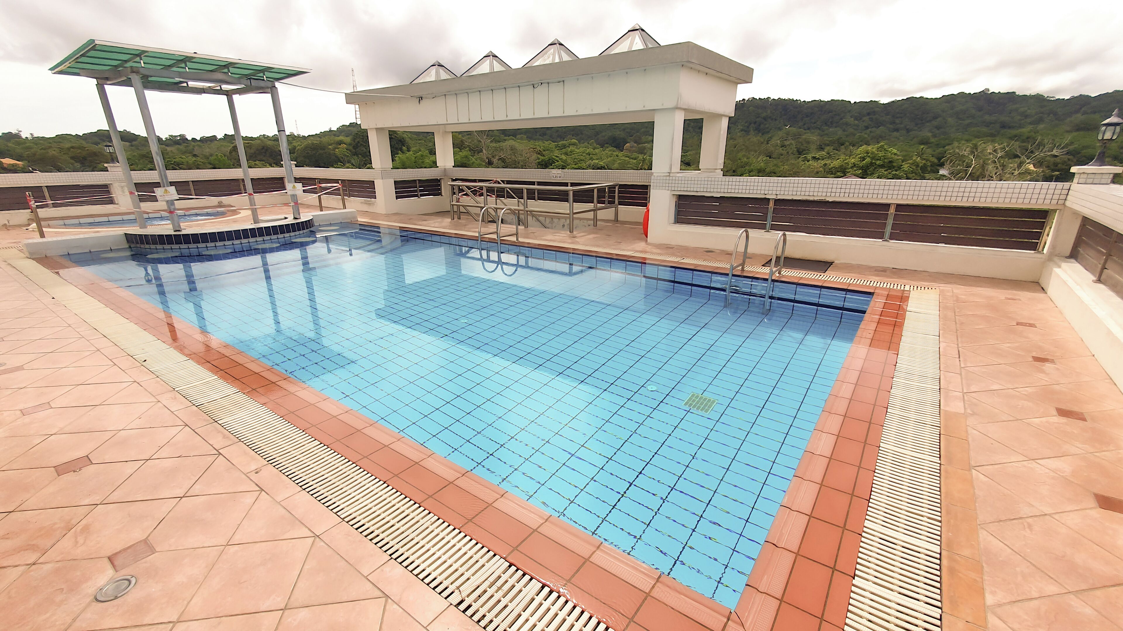A rectangular swimming pool with a tiled deck, a covered seating area, and a view of a forested hill in the background.