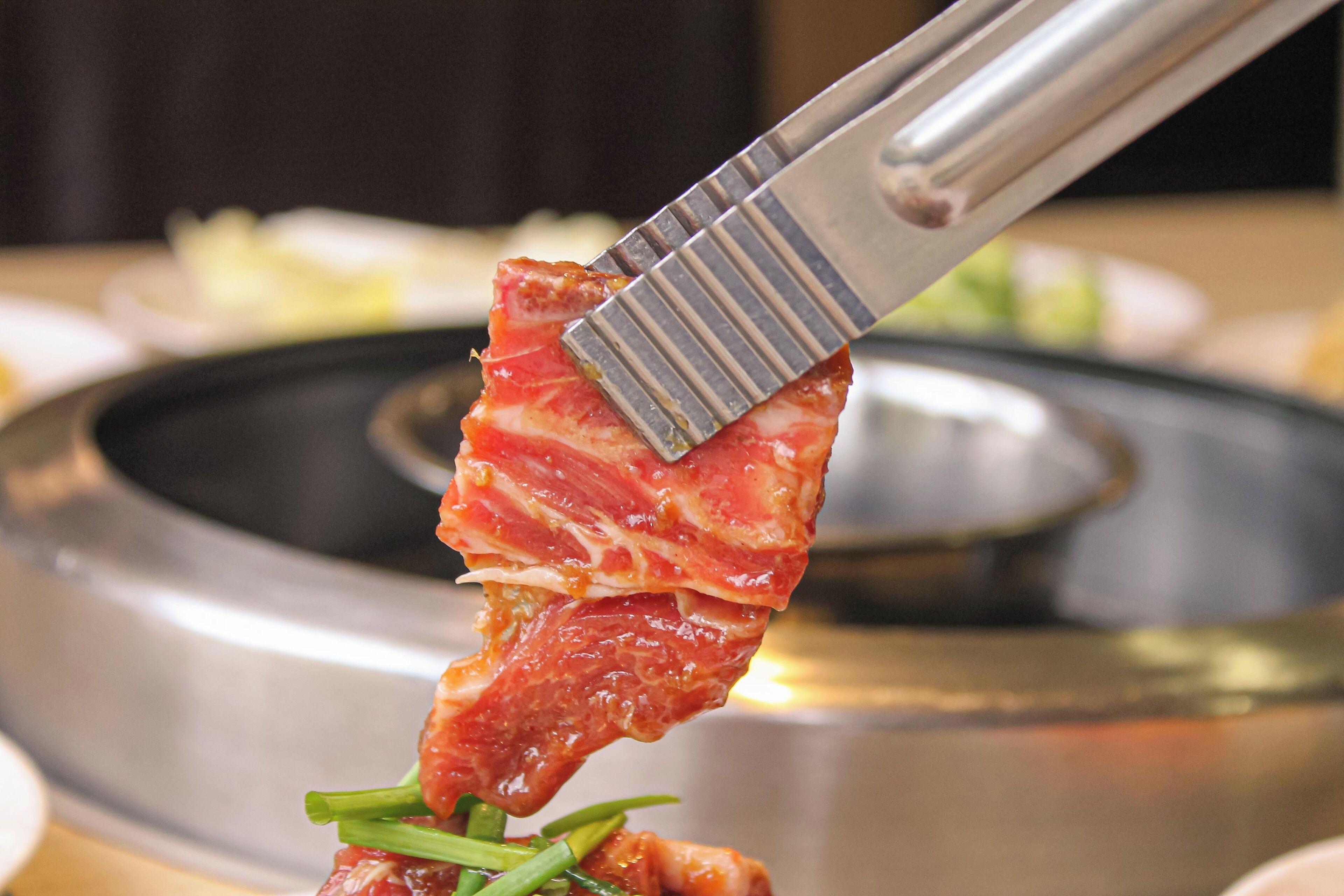 Un trozo de carne asándose sobre una superficie metálica con unas pinzas.