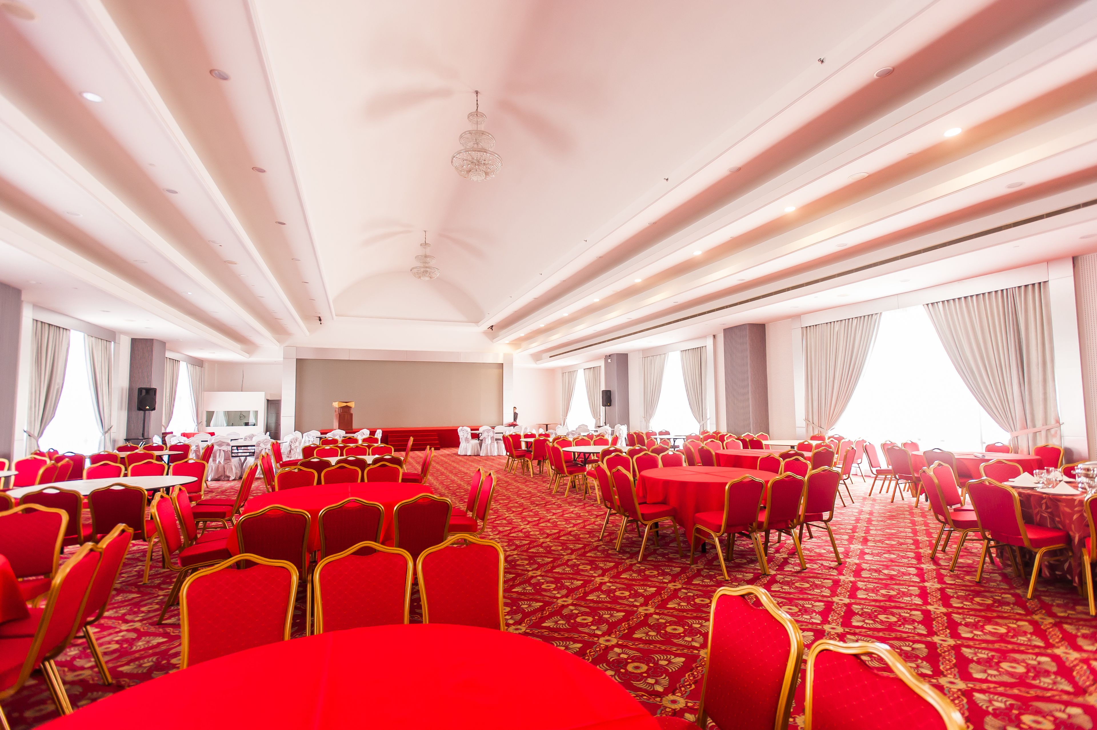 Un amplio salón de banquetes con mesas y sillas rojas, alfombra estampada y techo blanco con iluminación empotrada.