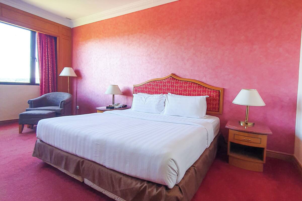 Una habitación de hotel con una cama grande, dos mesitas de noche con lámparas, una silla y un ventanal con cortinas.