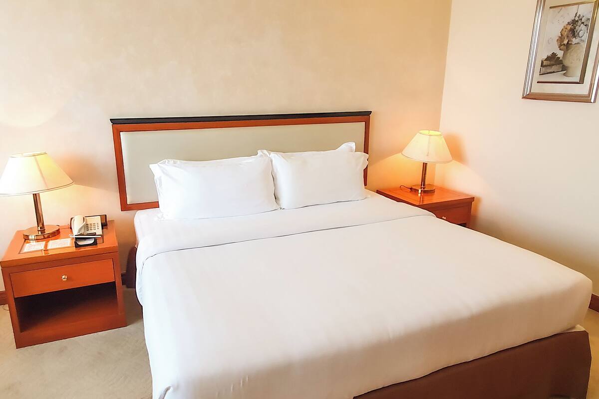 Una habitación de hotel con una cama grande, dos mesitas de noche con lámparas, un velador con teléfono y un cuadro en la pared.