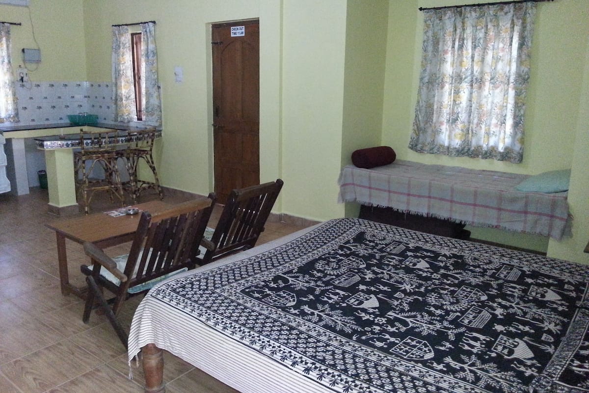 A room with a bed, a dining table with chairs, a kitchen area with a stove, and a window with curtains.