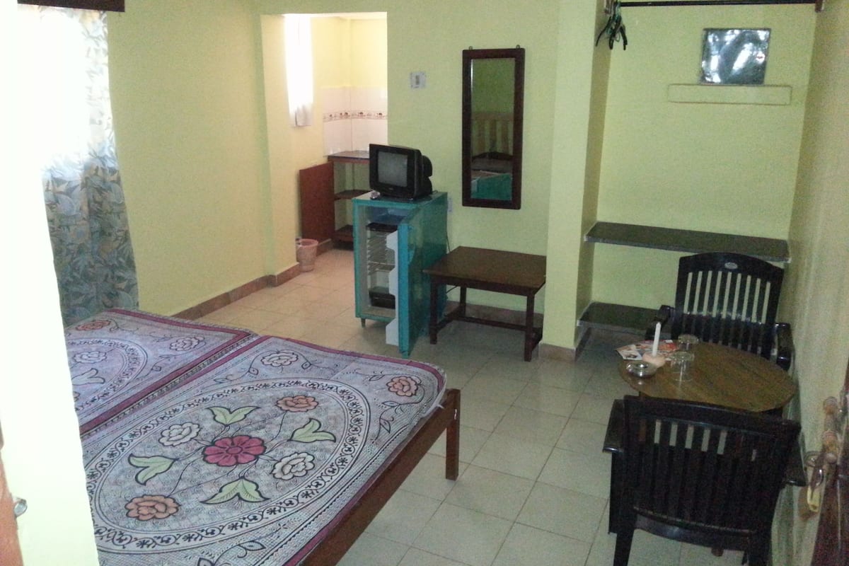 A room with a bed, a small table with a chair, a television on a stand, and a mirror on the wall.