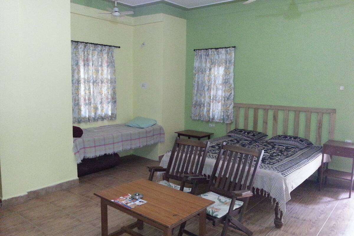 A small, simple room with a bed, a chair, a table, and a ceiling fan.