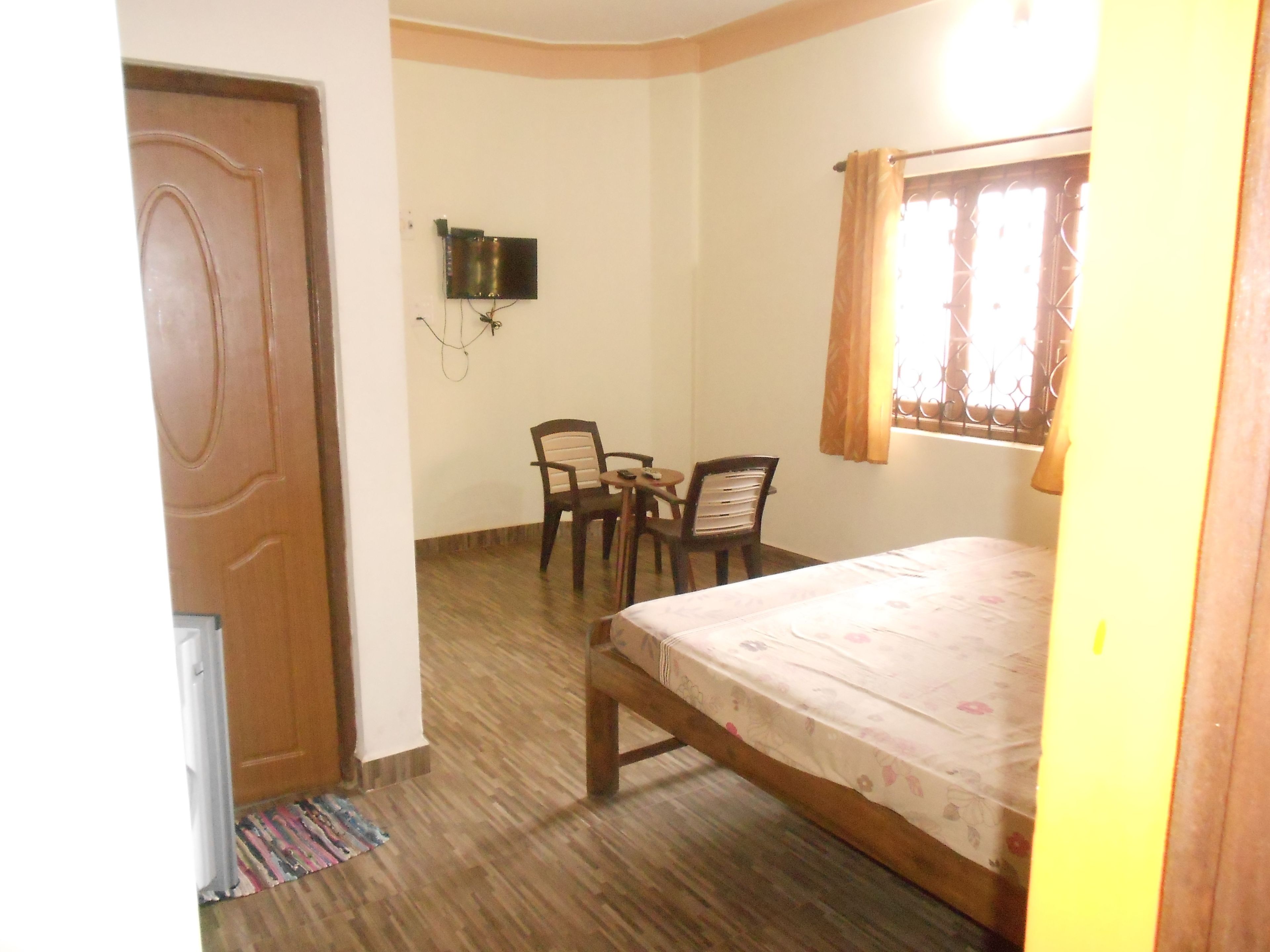 A small, simple room with a wooden door, a bed with a floral bedspread, a small table with chairs, a TV mounted on the wall, and a window with curtains.