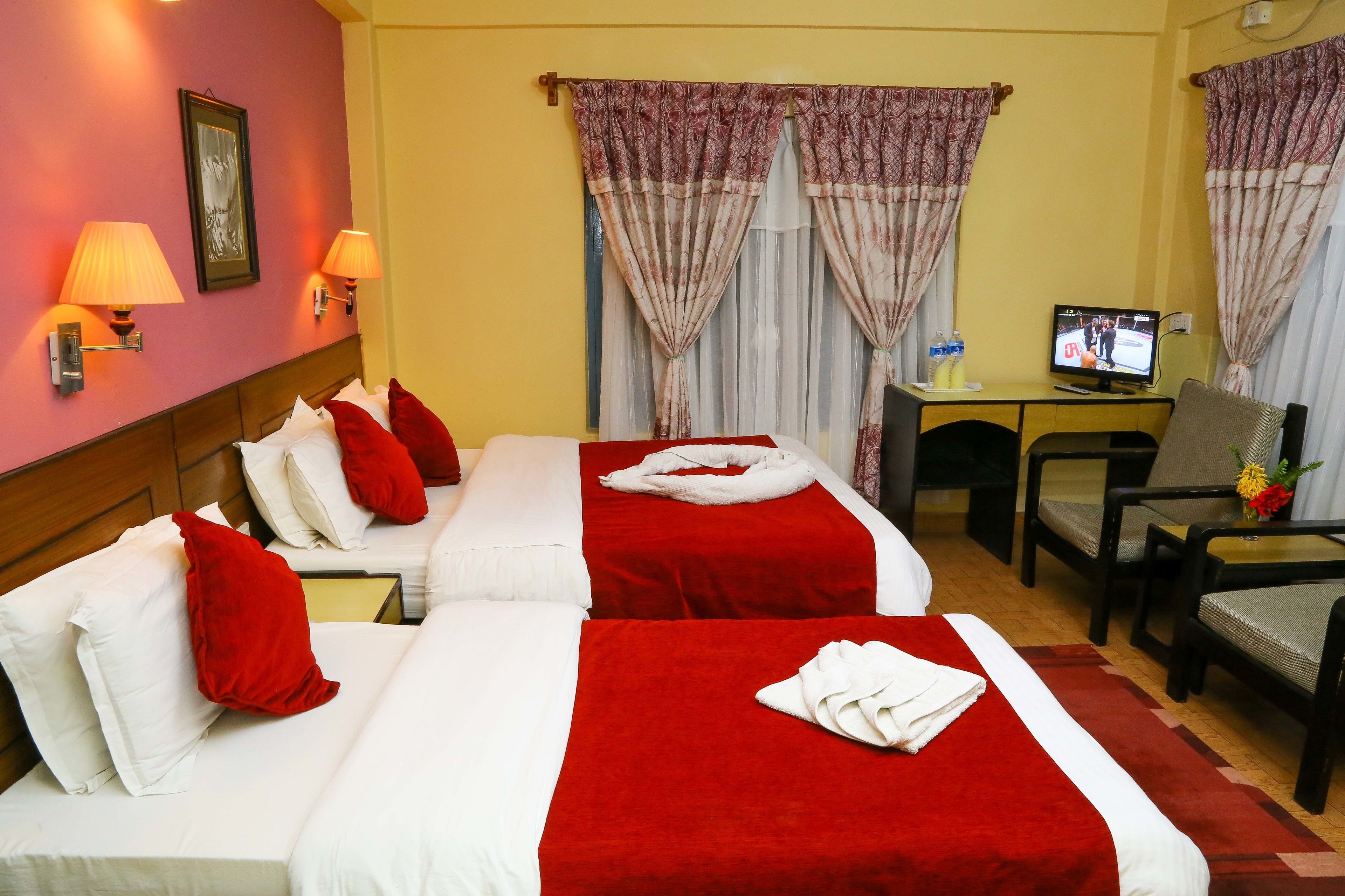 Camera d'albergo con due letti, biancheria rossa e bianca, una scrivania con televisore e un tavolino con un vaso di fiori.