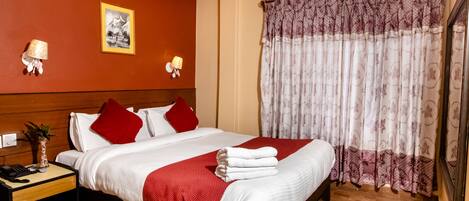 Habitación de hotel con cama, ropa de cama roja y blanca, mesita de noche con lámpara y ventana con cortinas estampadas.