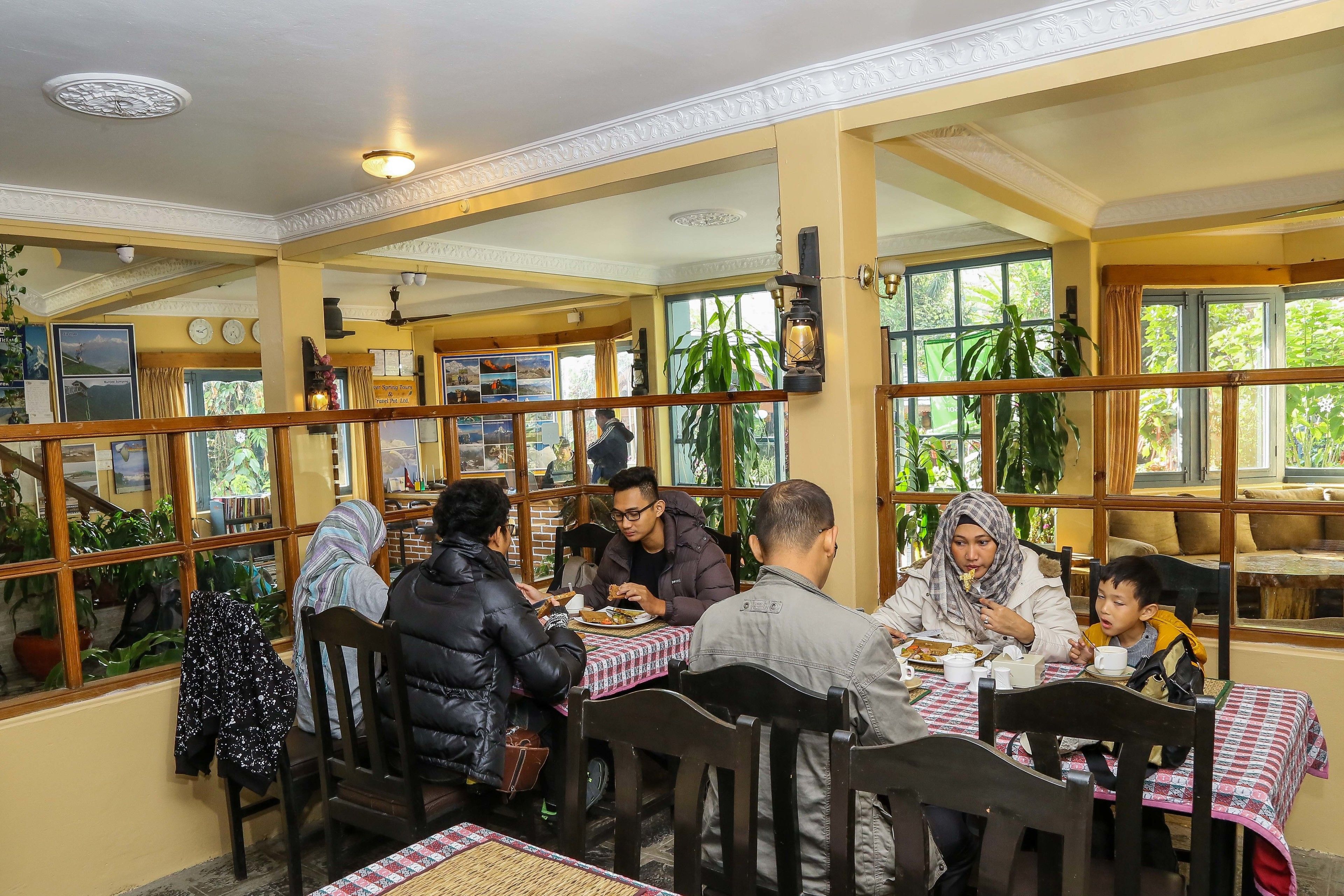 A restaurant with wooden tables and chairs, large windows with plants, and a traditional ceiling design.