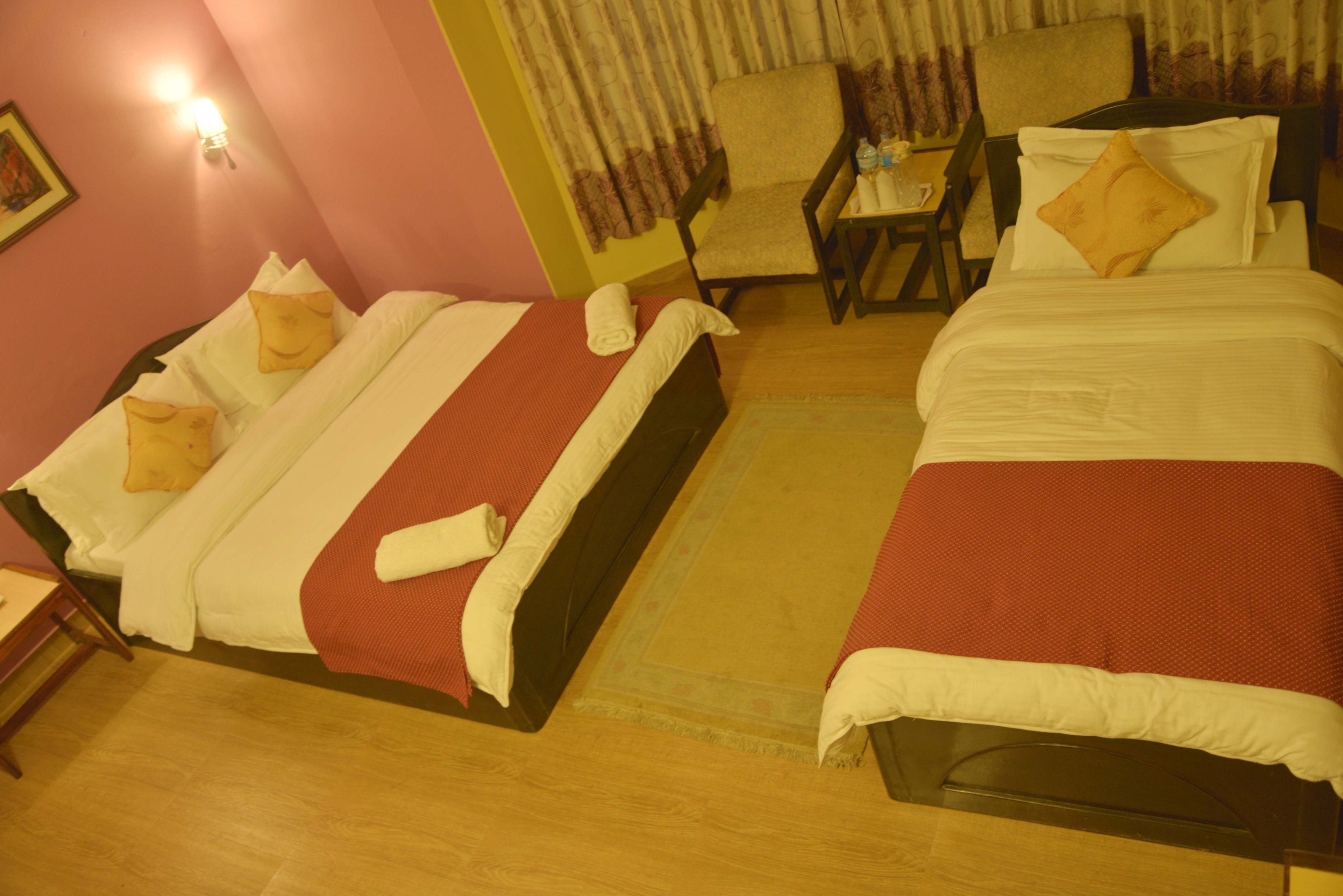 A hotel room with two beds, a chair, a small table, and a wall-mounted lamp.