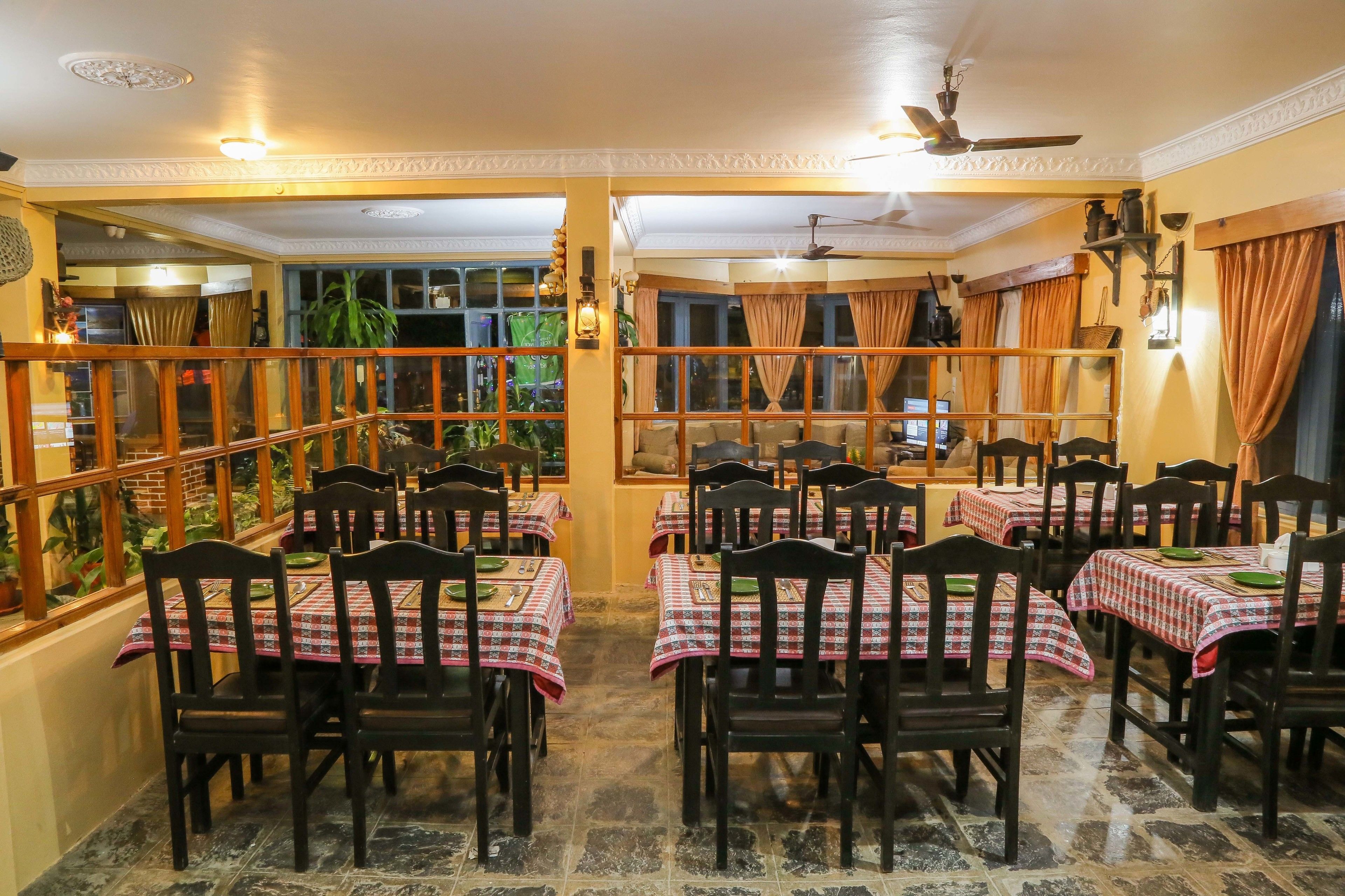 Un restaurante con varias mesas y sillas, cada una con mantel a cuadros. El suelo es de baldosas y hay ventanas grandes con cortinas.