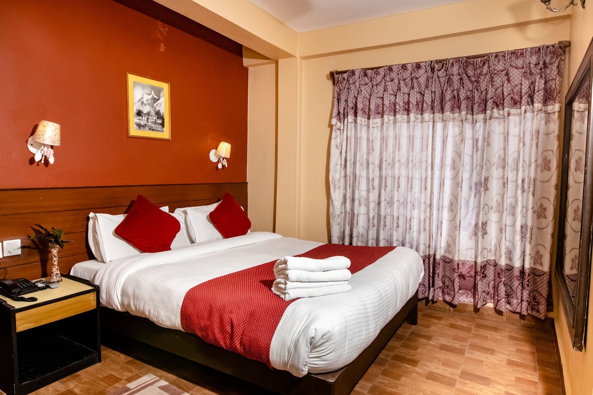 A hotel room with a bed, red and white bedding, a nightstand with a phone, and a window with patterned curtains.