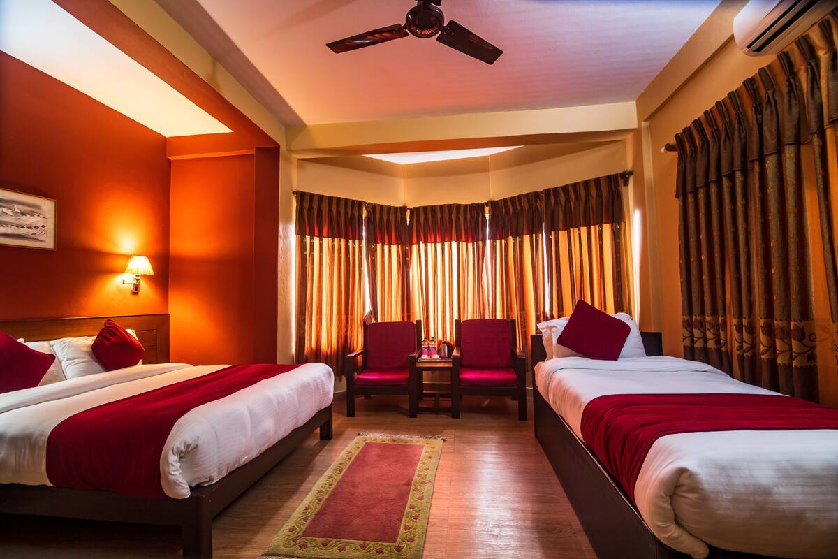 A hotel room with two beds, a ceiling fan, an air conditioning unit, a small table, and a window with curtains.