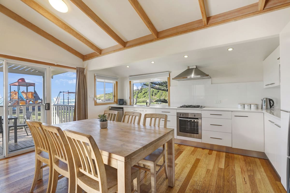 A modern kitchen with white cabinets, a stainless steel range hood, and a large window overlooking a playground.