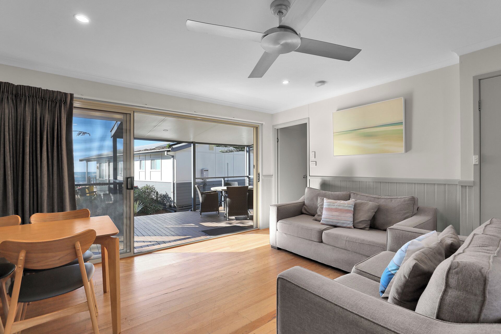 A modern living room with a grey sofa, a wooden dining table with chairs, a ceiling fan, and a sliding glass door leading to an outdoor area.