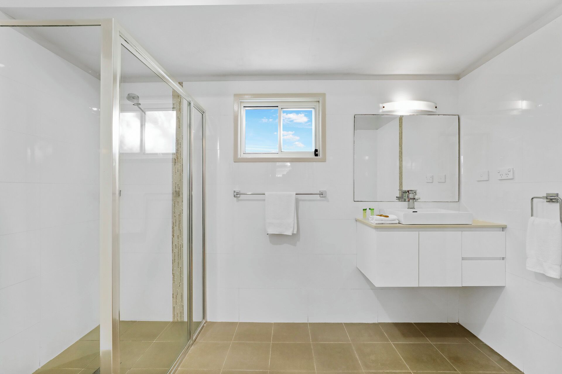 A modern bathroom with a glass shower enclosure, a window, a sink with a mirror, and a towel rack.