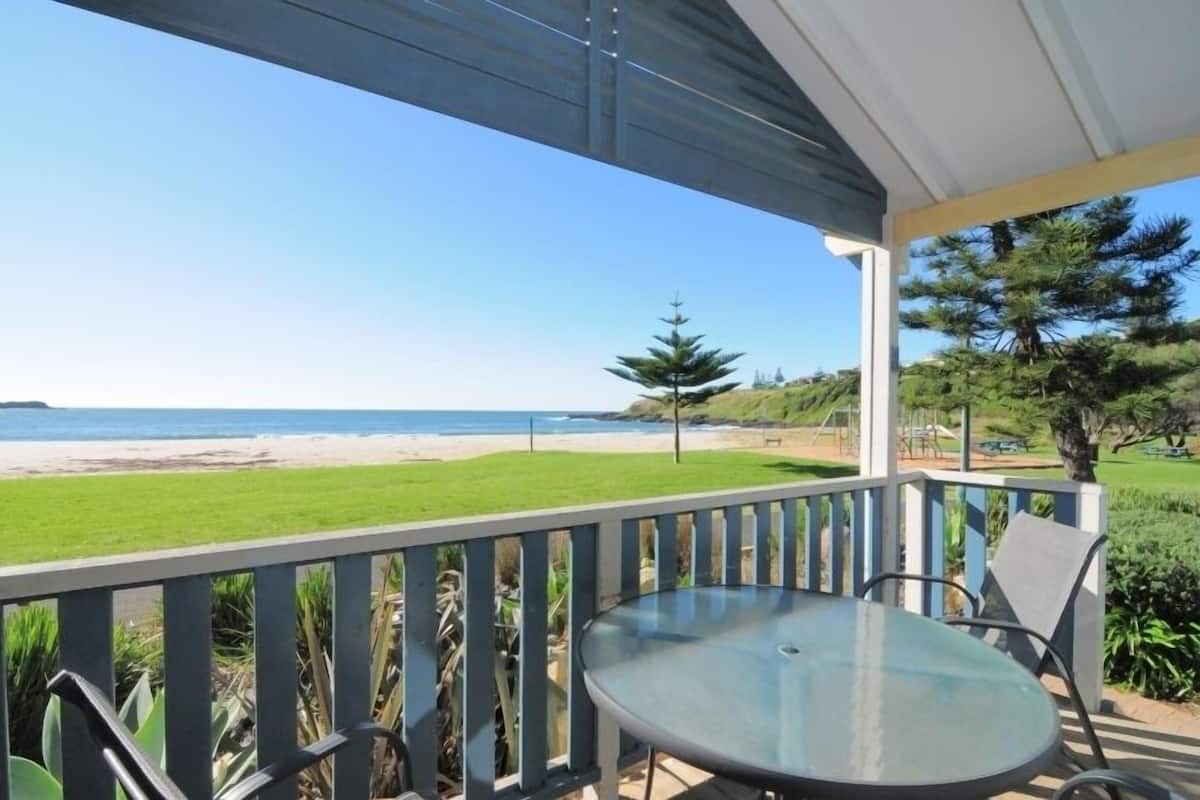 A balcony with a glass table and chairs overlooking a beach.