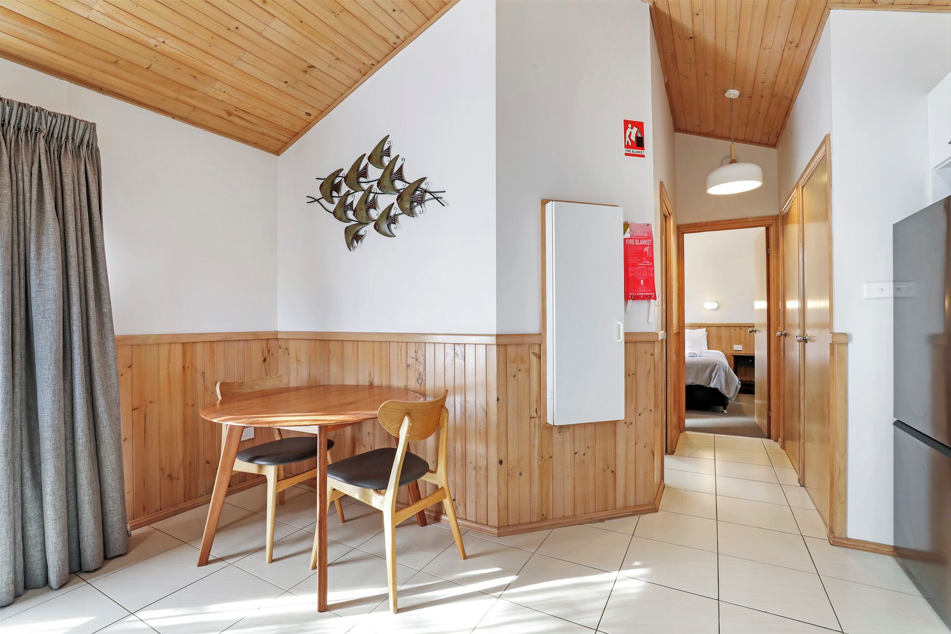 A room with a wooden ceiling, a dining table with chairs, a wall-mounted bird decoration, a whiteboard, and a door leading to another room.