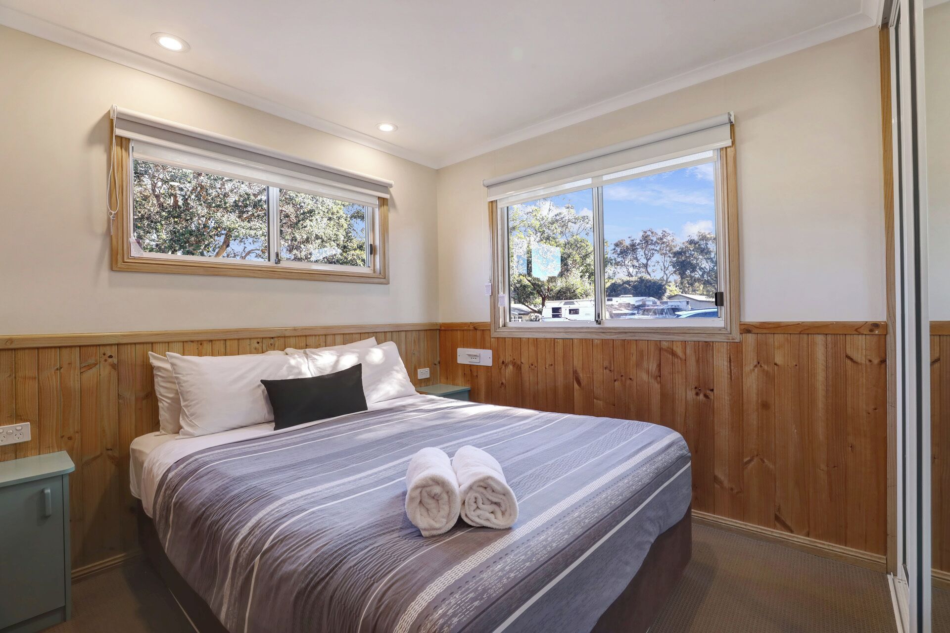 A bedroom with a bed, two windows, wooden paneling, and a view of trees and vehicles outside.