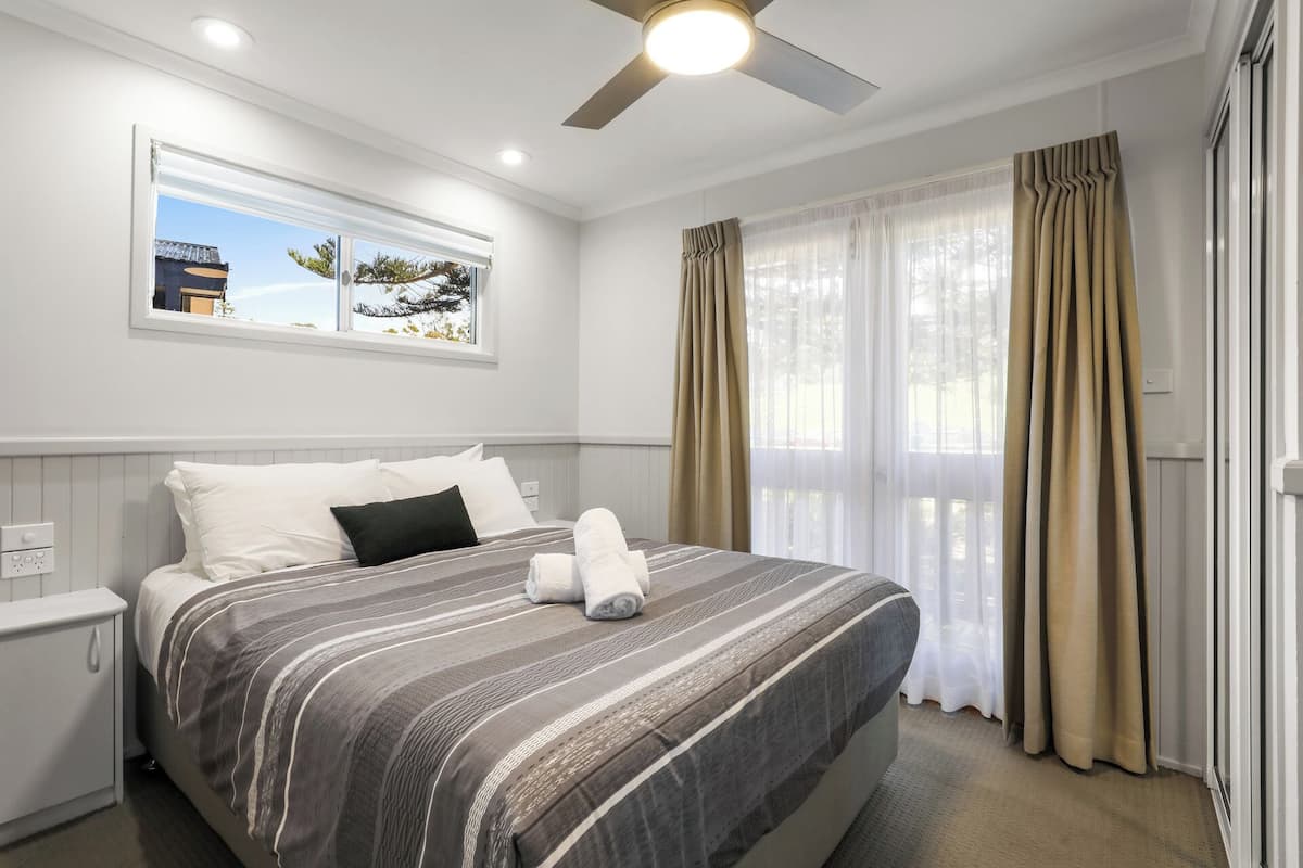 A hotel room with a bed, a nightstand, a ceiling fan, a window with curtains, and a view of the outdoors.