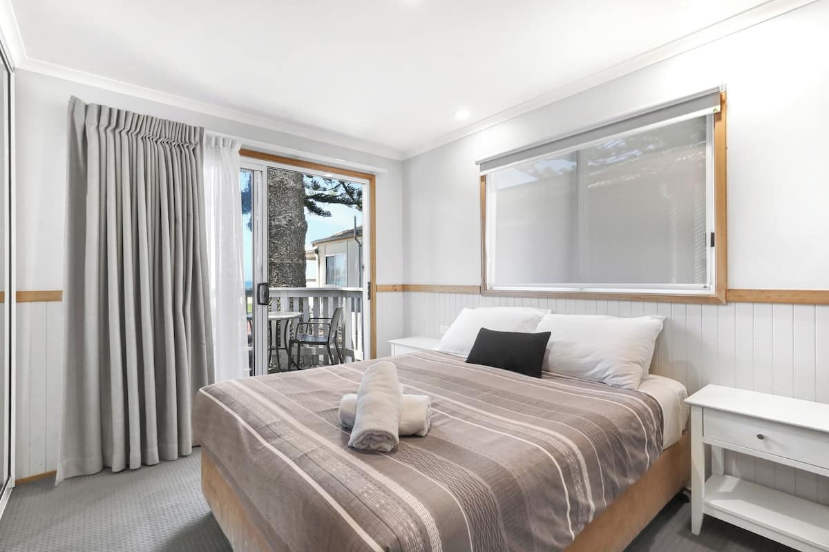 A bedroom with a bed, a nightstand, a window with blinds, and a view of the outside.