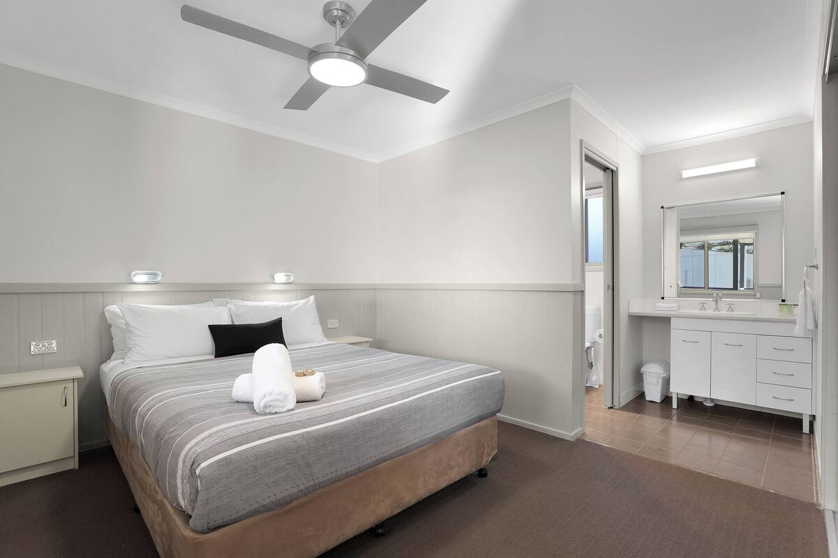 A modern bedroom with a bed, bedside tables, a ceiling fan, a bathroom with a sink and mirror.