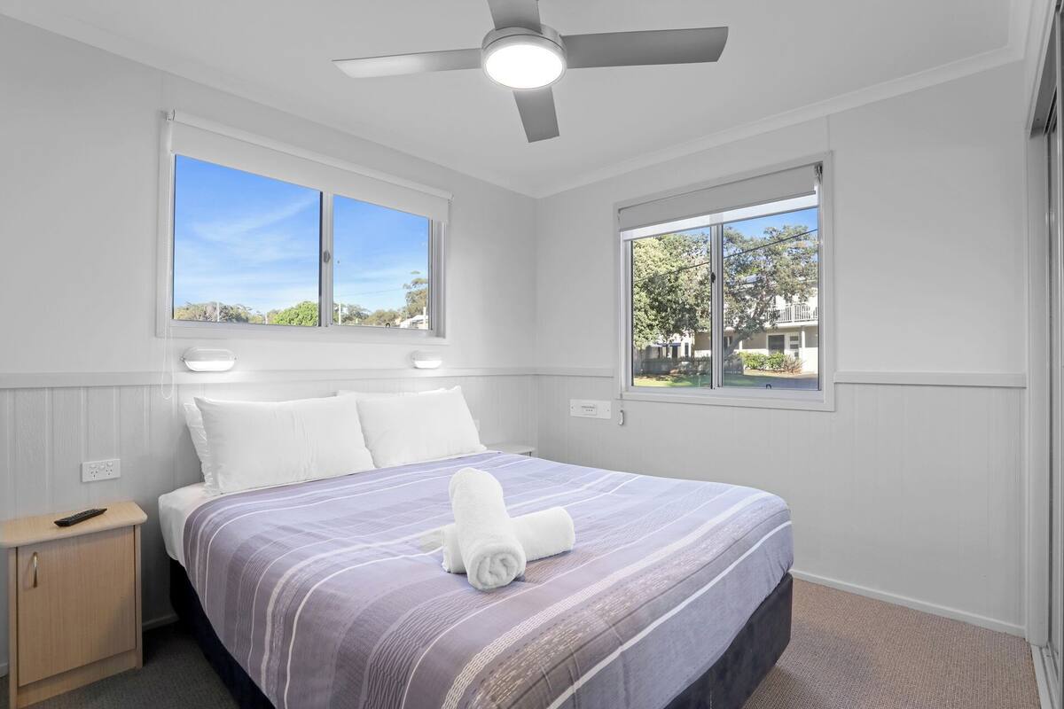 A bedroom with a bed, two windows, a ceiling fan, and a nightstand.