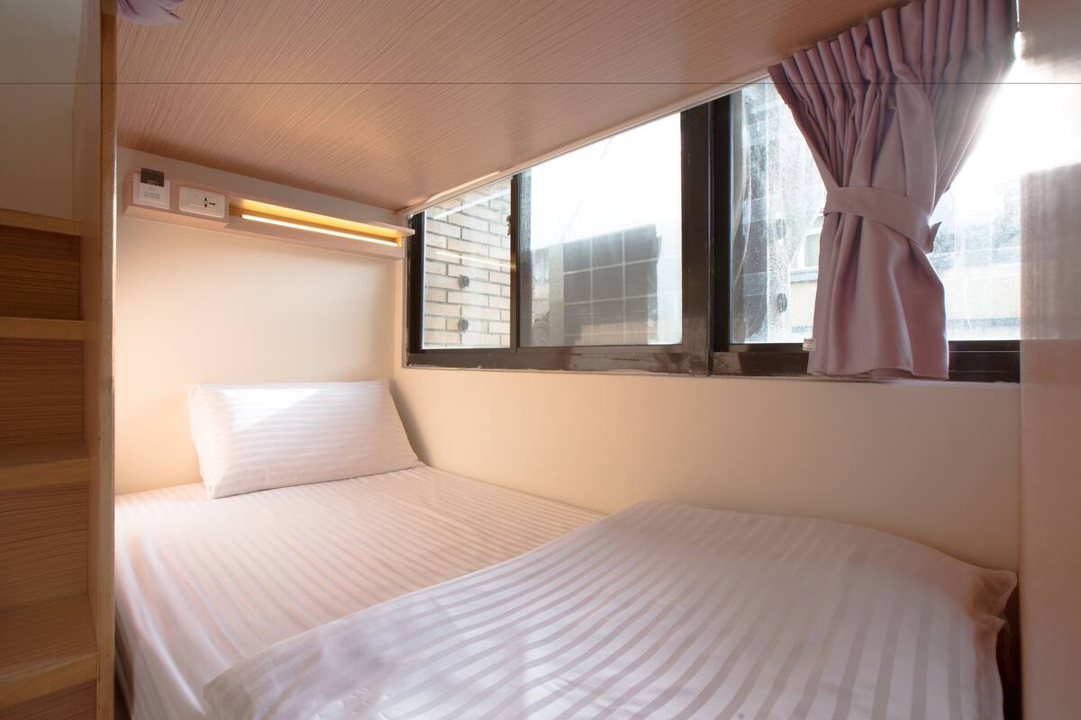 A small, well-lit room with two beds, a window with curtains, and a wooden shelf.