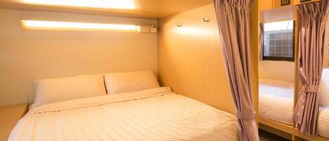 A single bed with a headboard, a bedside table, and a mirror. There are curtains on the windows and a ceiling light fixture.
