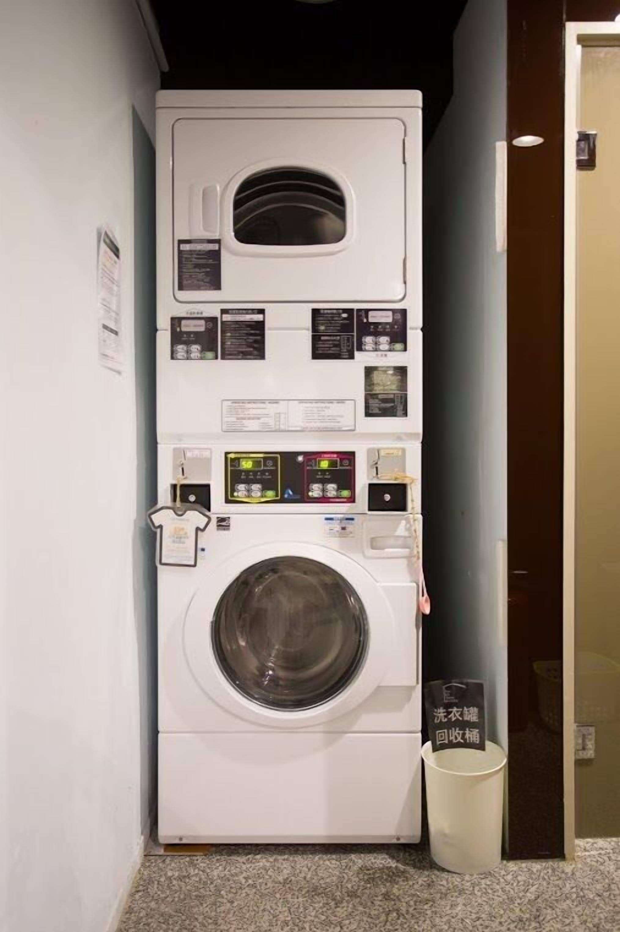 A compact, wall-mounted washing machine with a digital control panel, a front-loading drum, and a small trash bin beside it.