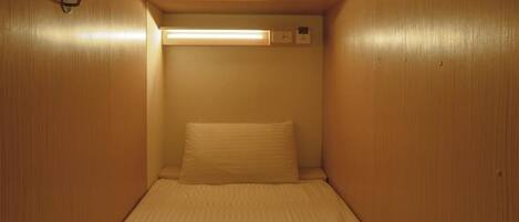 Una habitación pequeña y privada con una cama individual, una lámpara fijada a la pared y un termostato.