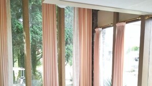 A large bed in a room with large windows and curtains, a view of greenery outside.