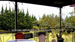 A covered outdoor area with a round table and wicker chairs, surrounded by greenery and a metal railing.