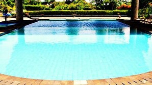 A large, clear swimming pool with a tiled edge, surrounded by a wooden deck and lush greenery in the background.