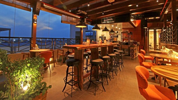 Un restaurante con zona de bar, techos altos y grandes ventanales con vistas a un paisaje urbano.