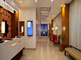 Um saguão de hotel moderno com uma mesa de recepção, uma tela digital, uma parede azul com painéis verticais e áreas de estar.