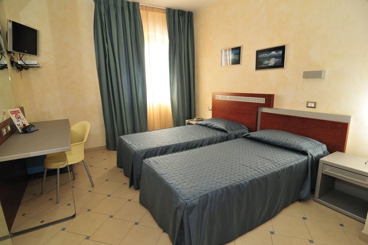 Une chambre d’hôtel avec deux lits simples, un bureau avec une chaise, un téléviseur fixé au mur et une fenêtre avec des rideaux.