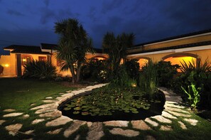 A well-lit garden with a pond, palm trees, and a pathway leading to a building with warm lighting. - La Locanda del Cavaliere (Latina)