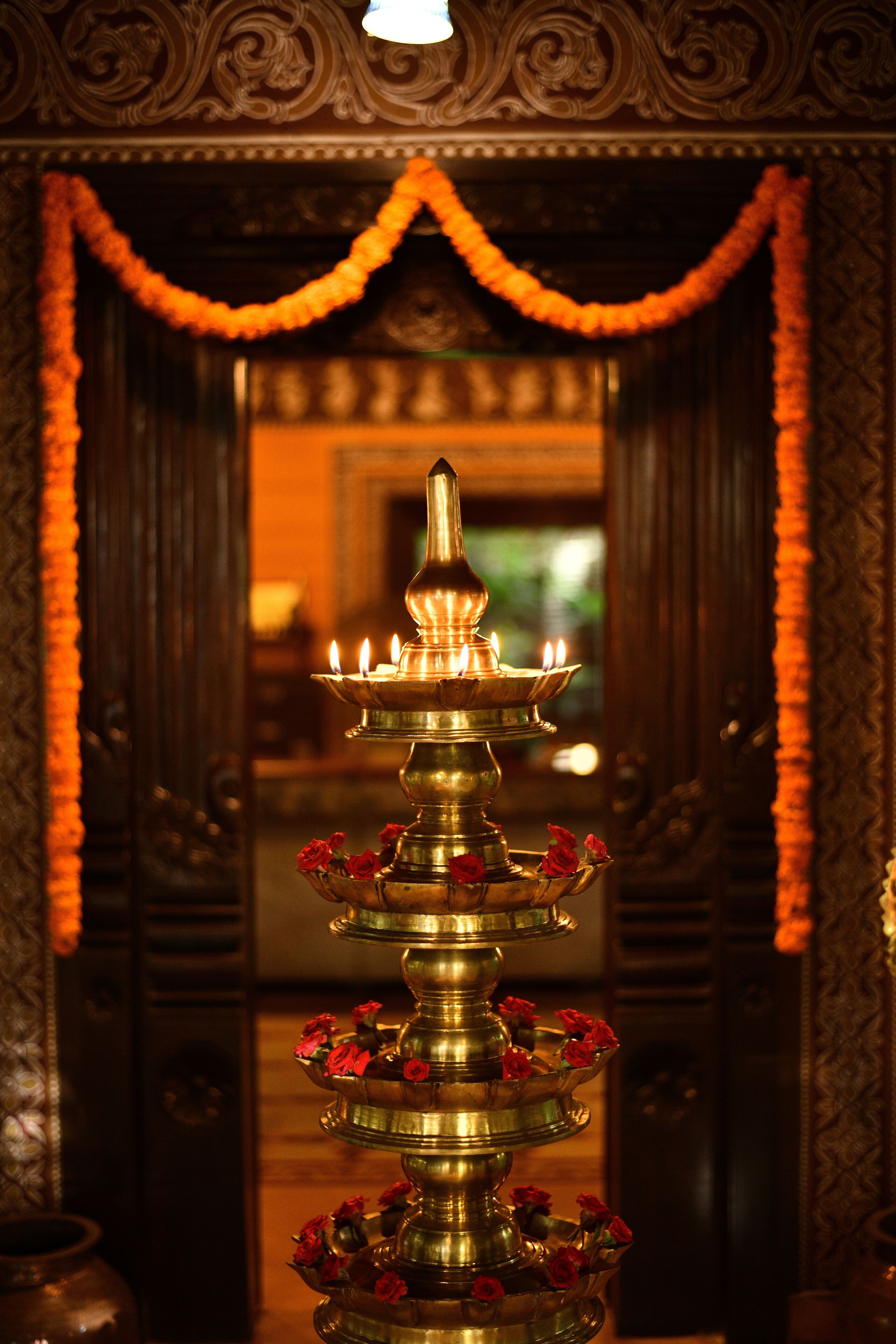 A traditional brass lamp with multiple tiers, adorned with red flowers, placed in a richly decorated room with ornate wooden paneling and a mirror reflecting the interior.